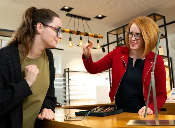Zwei Frauen probieren Brillen im Optikerladen aus und beraten sich freundlich am Tresen.