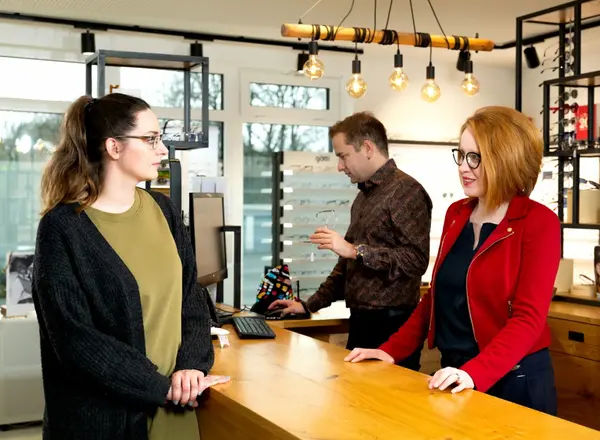 In einem modernen Optiker-Geschäft beraten zwei Mitarbeiter eine Kundin zu Brillenmodellen.