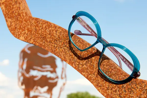 Blaue Brille aus Kunststoff liegt auf rostiger Metallskulptur vor blauem Himmel.