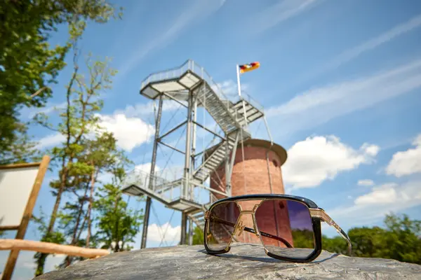 Eine Sonnenbrille liegt auf einem Stein vor einem Aussichtsturm mit deutscher Flagge.