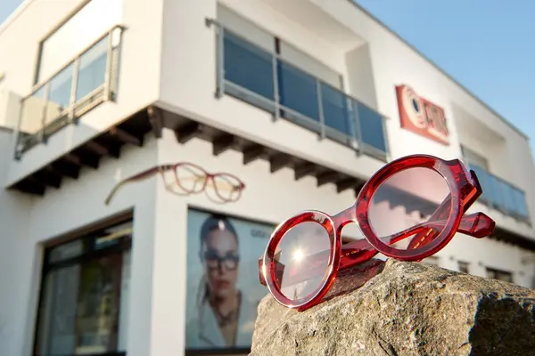 Rote Brille vor Optikergeschäft mit moderner Fassade und Werbeplakat für Brillenmode.