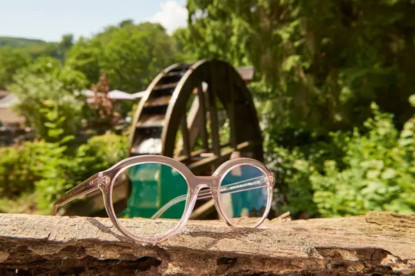 Transparente Brille aus Kunststoff liegt vor einem alten Wasserrad in grüner Natur.