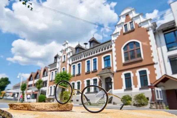 Runde Brille mit schwarzem Rahmen liegt vor historischem Altbau bei sonnigem Wetter.