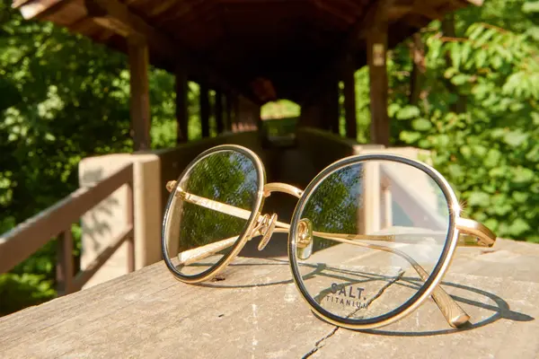 Goldene runde Brille aus Titan liegt auf Holzbank vor überdachter Holzbrücke im Grünen.