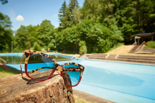 Sonnenbrille auf Baumstumpf vor leerem Freibad mit blauem Wasser und grünem Wald im Hintergrund.
